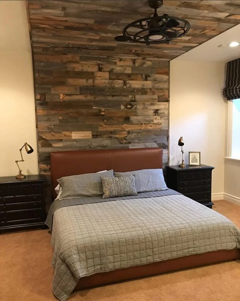 This bedroom is created a round a Feature Wall made of wood planks. The bed is covered with a grey duvet and grey pillows, accented by a deep brown headboard and black side tables.