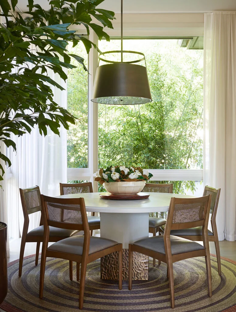 A small dining room with a white circular table topped with a flower arrangement. A plant is in the foreground.