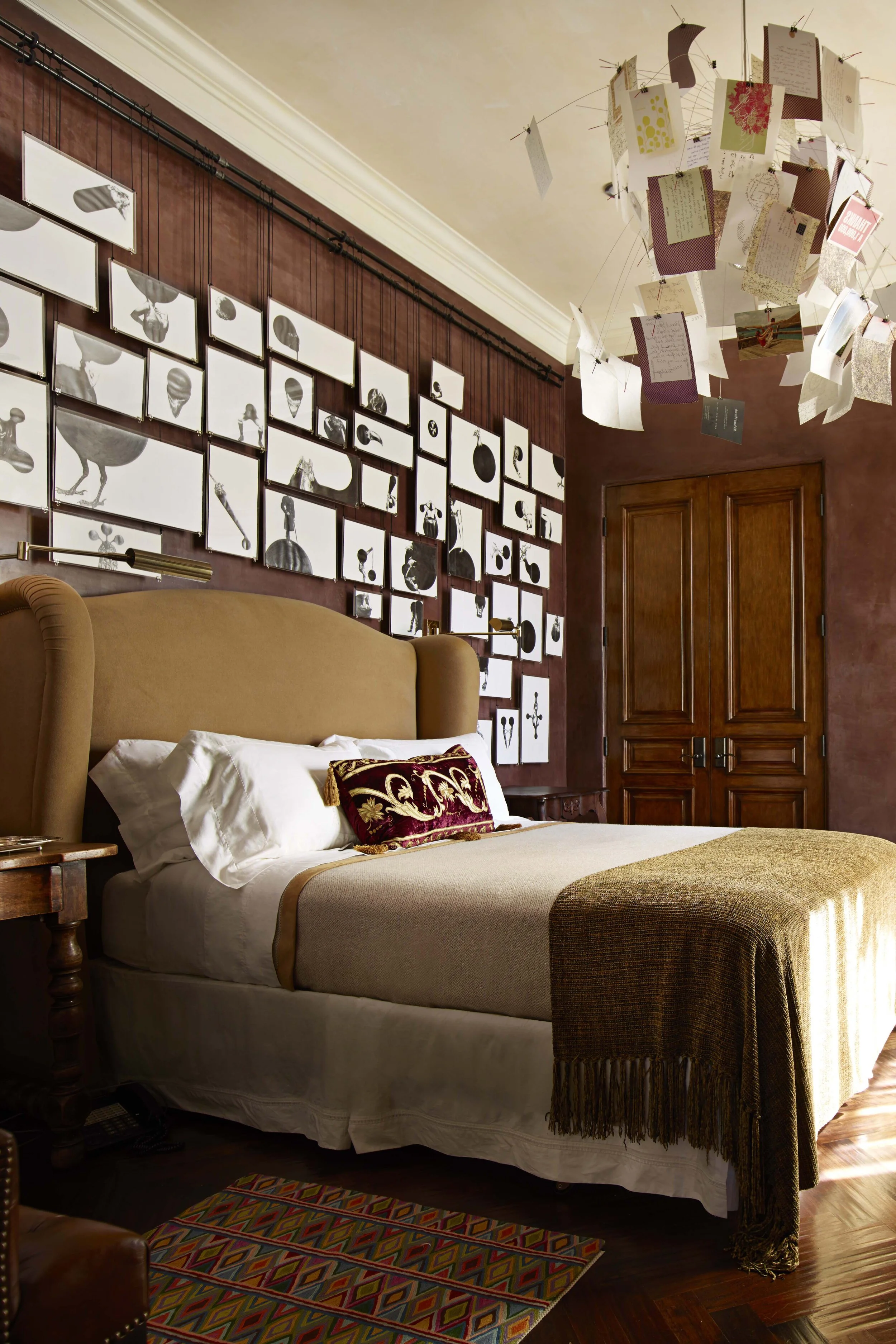 A lush bedroom with dark brown patina walls. Art is hung gallery style behind the bed. The chandelier is made of paper ephemera. Rehanging art is a great way to do a holiday refresh.