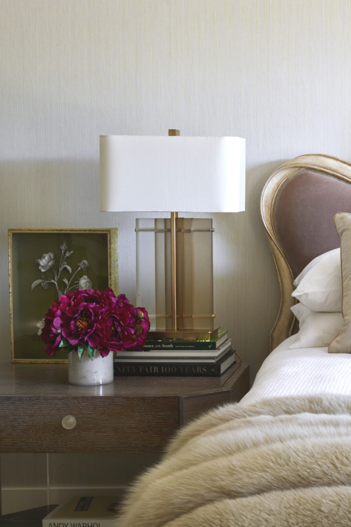 A night stand with a pot of fresh cut flowers, a modern lamp atop books, and a piece of artwork.